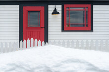 The Exterior Of A White House With A Vibrant Red Door, A Barn Light Fixture, Small Closed Glass Window With Red And Green Trim. There's A White Picket Fence In Front Of The Door And A Large Snowbank. 