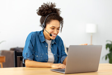 Successful African American woman in headset, office employee or call center worker sitting in a modern office talking, consulting client using video call, smile