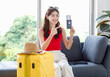 © Bangkok Click Studio - Asian young happy cheerful female traveler in casual summer outfit with sunglasses sitting smiling on cozy sofa in living room holding passport and boarding pass ticket with yellow trolley luggage