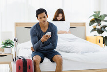 Asian Upset Stressed Depressed Frowning Face Young Husband Having Problem Fighting Arguing With Sad Wife Who Sitting On Bed. Male Packing Clothes And Stuffs In Trolley Luggage Ready To Leaving Home