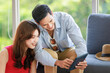 © Bangkok Click Studio - Asian traveler couple in casual outfit sunglasses and hat, husband smiling on sofa showing booking information in tablet computer to wife while helping packing clothes together in trolley luggage