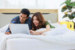 © Bangkok Click Studio - Asian happy romantic male female husband and wife lover couple lay down smiling together on pillow under blanket on bed using laptop notebook computer and credit card shopping making payment online