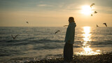 Fototapeta  - Caucasian woman feeding seagulls at sunset by the sea. 
