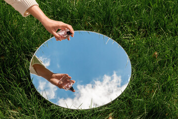 Wall Mural - perfumery and nature concept - hand with bottle of perfume and sky reflection in round mirror on summer field