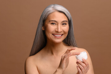 Poster - Enchanting topless middle aged Asian woman looks at the camera and laughing happily, holding jar of moisturizer cream isolated on brown background, charming korean lady advertising cosmetology product