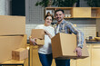 Portrait of young family husband and pregnant wife in new apartment holding cardboard boxes looking at camera and smiling