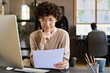 © pressmaster - Young manager in eyeglasses sitting at her workplace in front of computer and reading contract with serious expression