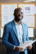 © pressmaster - Portrait of African successful businessman in blue suit holding documents and smiling at camera standing at office