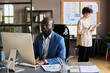 © pressmaster - African manager sitting at his workplace and concentrating on online work on computer with woman standing in background