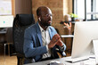 © pressmaster - African confident businessman in suit sitting on chair at his workplace in front of computer monitor and talking to colleague online