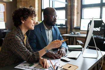 two multiethnic colleagues sitting at table in front of computer monitor and having online conferenc
