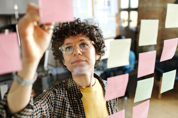 Young businesswoman in eyeglasses writing business plans on stickers hanging on glass wall during work at office