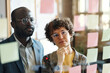 © pressmaster - Two employees in eyeglasses reading plans together on sticky notes hanging on glass wall at office