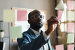 © pressmaster - Serious African businessman in suit and eyeglasses making notes on sticky notes hanging on glass wall at office