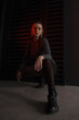 © NASTYA PALEHINA - teen fashion. young brunette girl in a shiny silver bodysuit and boots is sitting fashion on floor with hands on the knees near black iron wall background with red light. fashion concept, free space