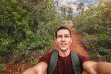 Young Happy Man Takes Selfie Photo During His Awesome Hiking Trek Somewhere In Africa National Park, Walking By Red Soil Footpath In Jungle