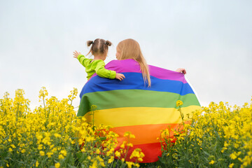 A woman with a little child in arms and with a bright rainbow unfurled LGBT flag in a field with rapeseed flowers against the sky. The concept of family well-being in homosexual families