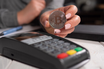 Close-up of a young woman holding a silver bitcoin coin near the terminal. Pay for purchases with cryptocurrencies. BTC.