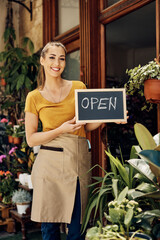 Wall Mural - Young happy florist holding open sign at her flower shop and looking at camera.