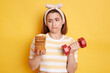 © sementsova321 - Indoor shot of upset pensive woman wearing t shirt and hair band posing isolated on yellow background, holding dumbbell in one hand and looking at sweet cookie in another hand.
