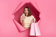 © sementsova321 - Image of excited surprised woman wearing striped shirt posing in breakthrough of pink background, standing with shopping bags and smart phone, checking cashback.