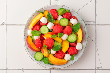 Wall Mural - Summer fruit salad with mozzarella cheese, cucumber and mint in a plate on white tile background. top view