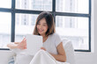 © kaew6566 - Positivity asian woman holding digital tablet sitting with pillow white bed beside window at home. Happy young female sitting relax looking something on tablet social media at home. Technology concept