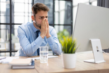 Unhappy frustrated young male holding head by hands sitting with computer behind desk at office.