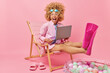 © wayhome.studio - Joyful surprised curly woman dreams about summer vacation wears goggles shirt and flippers works on laptop computer poses on deck chair against pink background. People holidays tourism concept