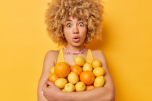 Impressed Shocked Woman With Curly Blonde Hair Embraces Heap Of Juicy Fresh Oranges And Lemons Keeps Mouth Opened From Wonder Isolated Over Vivid Yellow Background. Citrus Fruits Full Of Vitamins