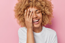 Headshot Of Positive Carefree Woman Makes Face Palm Smiles Toothily Keeps Eyes Closed Has Pleasant Feelings Dressed In White T Shirt Isolated Over Pink Background. People And Emotions Concept