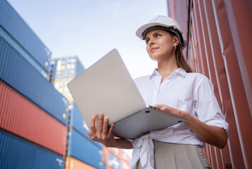 Dock manager or engineer worker in causual suit standing in shipping container yard holding laptop with smile. Import and export product. Manufacturing transportation and global business concept.