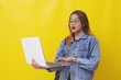© ArvStd - Amazed asian young college student standing while holding a laptop. Isolated on yellow background