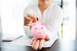 © OkayStocker - female calculating expenses and having ideas for saving money, Her hand was holding the piggy bank and was picking up the coin, saving money or savings concept.