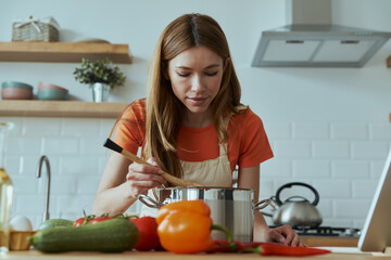 Wall Mural - Confident young woman cooking soup while standing at the domestic kitchen