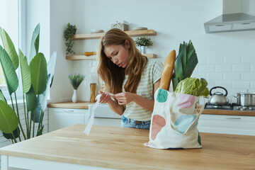 Wall Mural - Concentrated young woman checking receipts while standing at the domestic kitchen