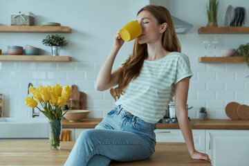 Wall Mural - Beautiful young woman drinking coffee from yellow cup while sitting on the kitchen counter