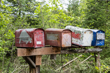 Closeup Selective Focus View Of Four Old Rusty Mailboxes On A Totting Wooden Post With Blurry Trees In Background In Rural Village. Copy Space Above.
