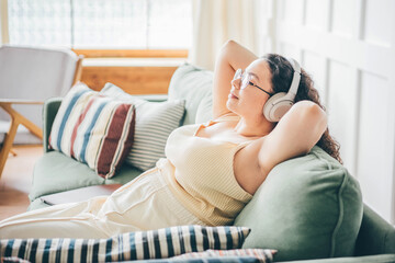 Wall Mural - Curly haired overweight young woman wearing comfortable clothes lies on green sofa listening to music in wireless headphones in stylish room