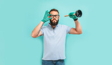 Young Handsome Man Wearing Plumber Uniform Holding Toilet Plunger Looks Happy. Professional Cleaning Of Clogged Pipes. Studio Shot On Blue Background. Funny Promotion Poster