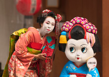 Kyoto, Japan - March 26 2020: Japanese Female Posing In A Maiko's Outfit Aside A Character Depicting A Maiko With A Speech Balloon Where Is Wrote The Hashtag Maiko Experience.