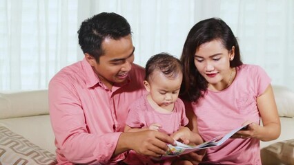 Wall Mural - Baby girl reads storybook with her parents at home