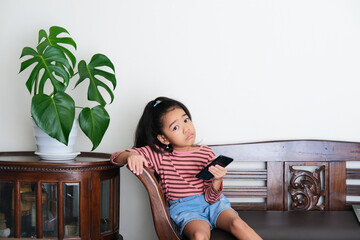 Wall Mural - Asian little kid sitting relax in a couch while holding mobile phone and showing confused expression