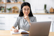 © kucherav - Happy asian female student with headset looking at laptop screen, studying remotely using laptop, taking notes on notepad sitting from home, e-learning concept
