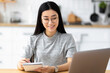 © kucherav - Focused asian female student studying remotely using a laptop, taking notes on notepad sitting frome home during online lesson, e-learning concept