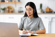 © kucherav - Focused asian female student with headset studying remotely using a laptop, taking notes on notepad sitting frome home during online lesson, e-learning concept