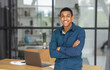 © kucherav - Portrait of successful young african american businessman with arms crossed standing in modern office. Happy entrepreneur looking at the camera and smiling friendly