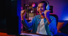 Happy Asia Guy Gamer Wear Headphone Participation Play Video Game Colorful Neon Lights Computer In Living Room At Night Modern House. Esport Streaming Game Online, Home Quarantine Activity Concept.