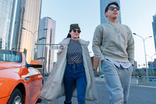 A Fashionable Young Couple With Sunglasses And Walking Beside An Orange Luxury Sports Car