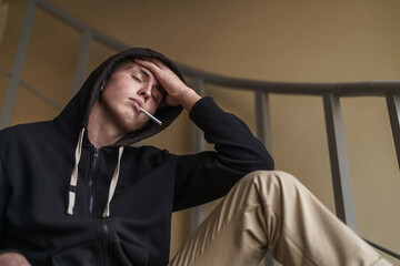 Wall Mural - frustrated young man in a hood and with a cigarette in his mouth is sitting on the stairs and looking away. Teenage transition age. The concept of loneliness and problems.